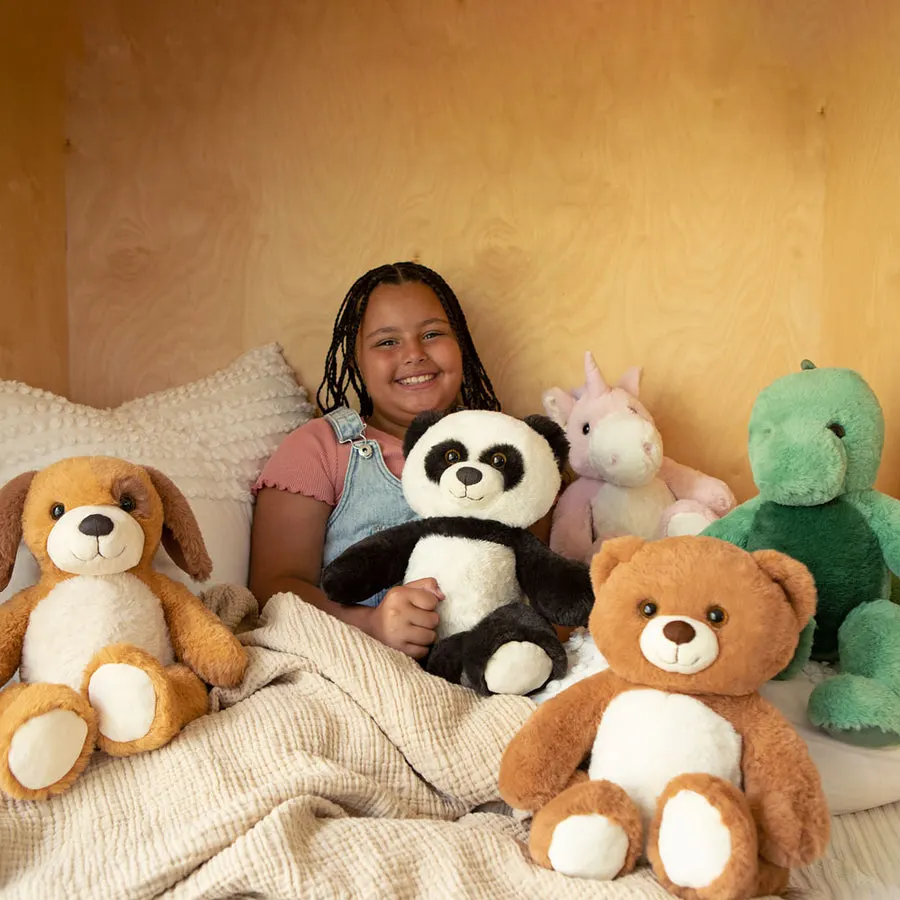 A smiling child sits in bed under a cream blanket, surrounded by an assortment of plush animals including a panda, a brown teddy bear, a dog, a pink unicorn, and a green dinosaur.