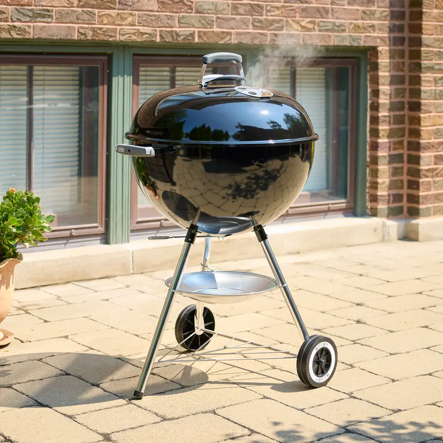 A black Weber kettle charcoal grill with a wisp of smoke rising from the lid, standing on a sunlit brick patio in front of a stone-and-brick building with green-trimmed windows.