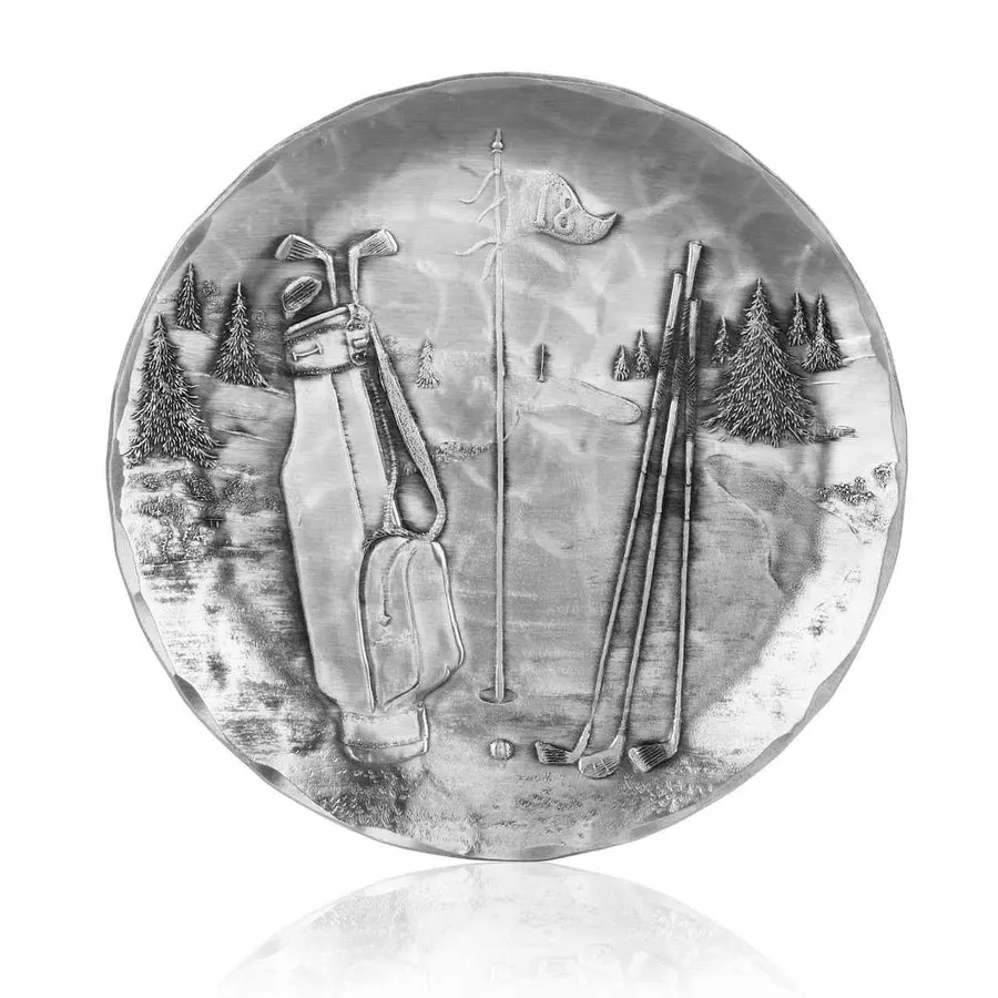 A round hand-hammered aluminum coaster embossed with a golf scene showing a golf bag, clubs, a flagstick, and pine trees.
