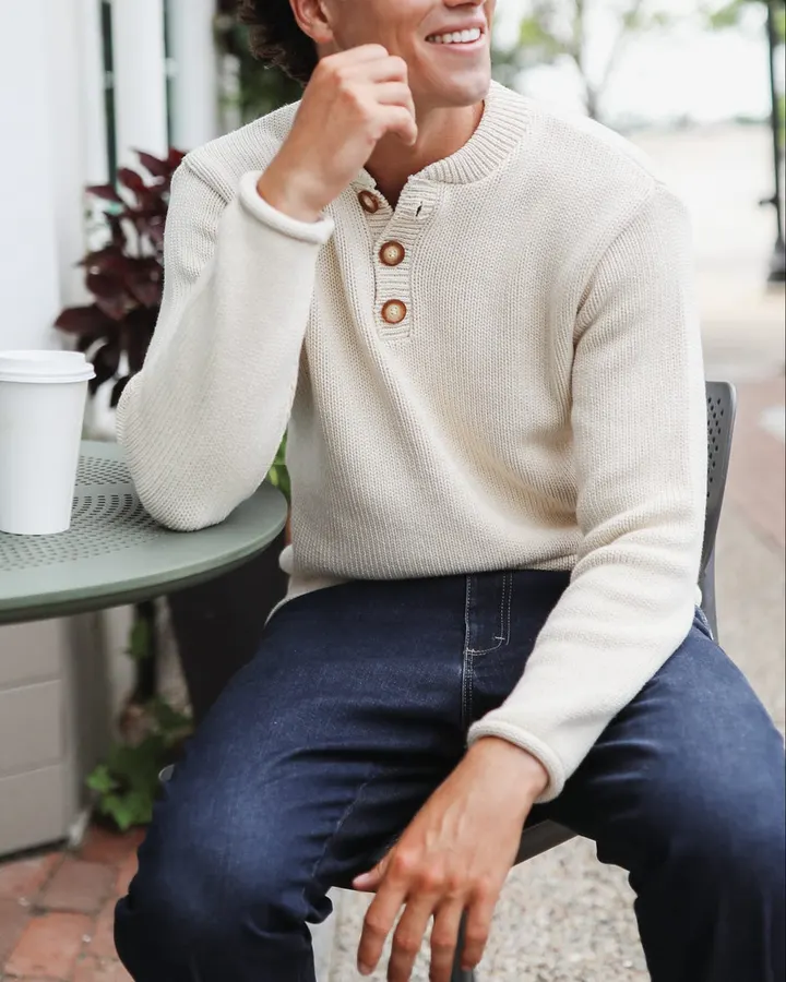A smiling man in a cream three-button henley sweater sits at an outdoor cafe table with a paper coffee cup, one hand raised near his chin.