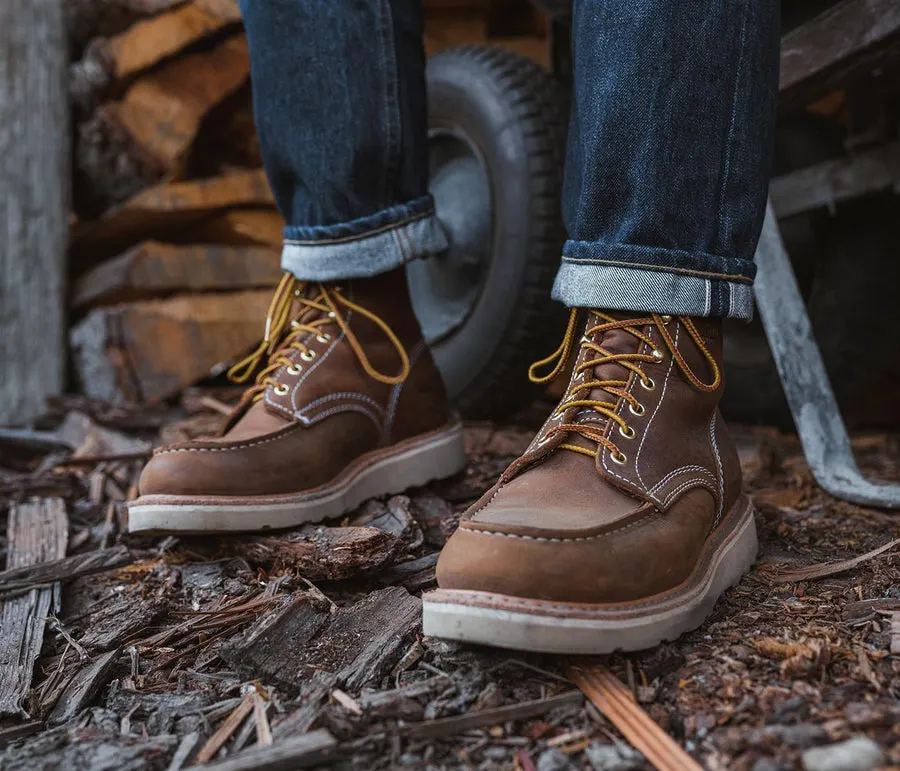 A person's feet wearing brown leather moc-toe work boots with gold laces and cream soles, standing on wood chips and bark mulch in front of a woodpile and a wheelbarrow tire.