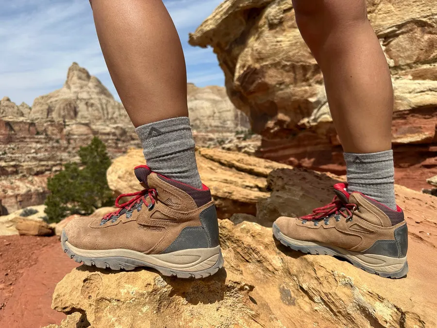 A hiker's lower legs in grey wool hiking socks and tan-and-red leather hiking boots, standing on red sandstone rock with a desert canyon landscape in the background.