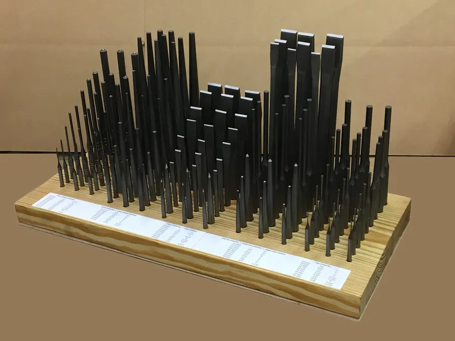 Dozens of black steel punches and chisels of varying lengths stand upright in a wooden display board, photographed against a plain tan background.