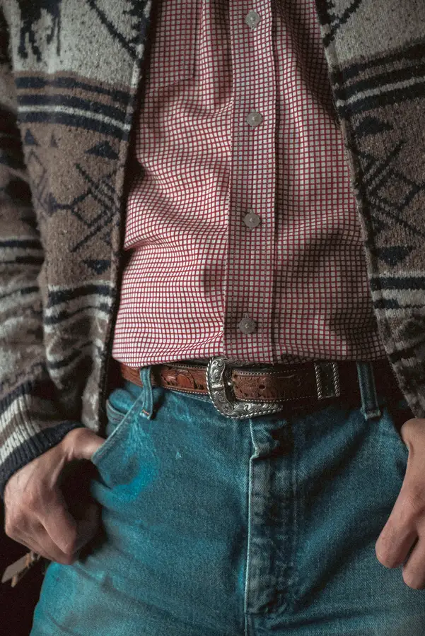 Close crop of a person's waist wearing faded jeans and an open patterned cardigan over a red gingham shirt, cinched with a brown tooled-leather belt and an engraved silver buckle.