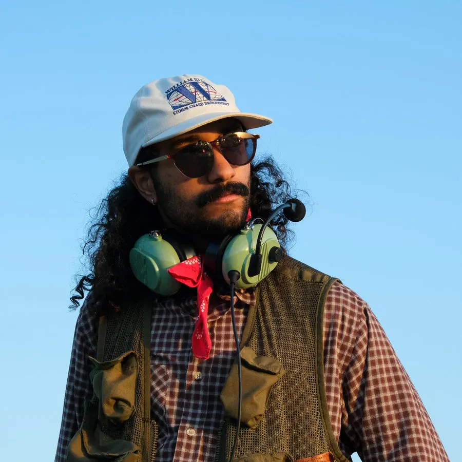 A man with long curly hair and a mustache wears a white William Ellery Storm Chase Department cap, sunglasses, and a plaid shirt under a mesh vest with green headphones around his neck, photographed outdoors against a clear blue sky.