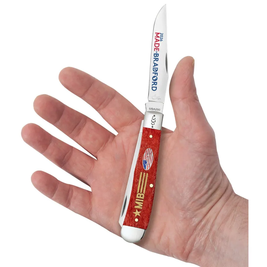 A hand holding open a red-scaled Case trapper knife with 'Made in Bradford' and 'Celebration' etched on the blades, palm and fingers framing the knife against a white background.